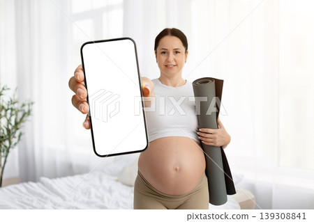 Pregnant woman holding yoga mat under arm and showing blank smartphone screen Pregnant woman holding yoga mat under arm and showing blank smartphone screen 139308841