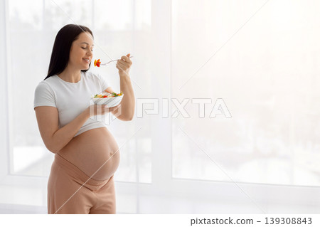 Pregnant woman standing near bright window eating fresh vegetable salad Pregnant woman standing near bright window eating fresh vegetable salad 139308843