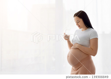 Pregnant woman standing near window holding glass of water and touching belly Pregnant woman standing near window holding glass of water and touching belly 139308849