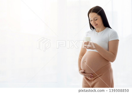 Pregnant woman standing near window holding glass of milk and cradling belly Pregnant woman standing near window holding glass of milk and cradling belly 139308850
