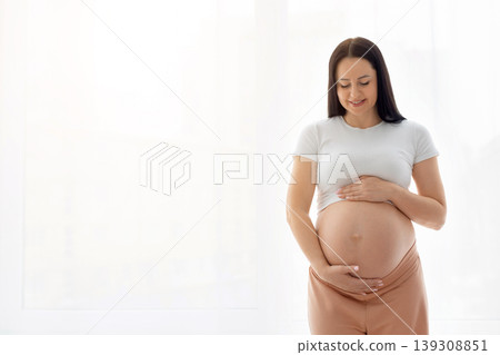 Smiling pregnant woman standing near window tenderly holding belly with both hands Smiling pregnant woman standing near window tenderly holding belly with both hands 139308851