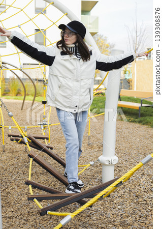 Young woman balancing on a rope bridge in an outdoor playground, expressing playful movement and childlike energy. Young woman balancing on a rope bridge in an outdoor playground, expressing playful movement and childlike energy. 139308876