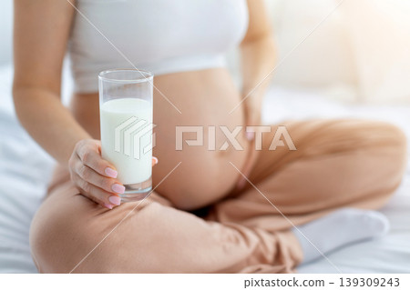 Pregnant woman sitting on bed holding glass of milk beside belly Pregnant woman sitting on bed holding glass of milk beside belly 139309243