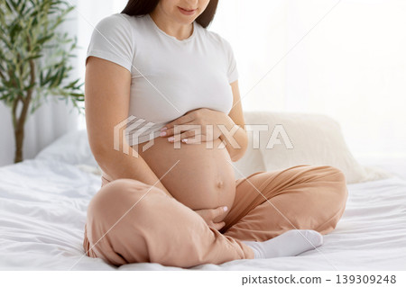 Closeup of pregnant woman sitting cross legged on bed tenderly holding belly Closeup of pregnant woman sitting cross legged on bed tenderly holding belly 139309248