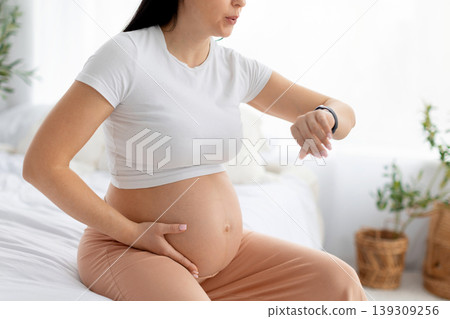 Pregnant woman sitting on bed checking smartwatch while holding belly, timing contractions 139309256