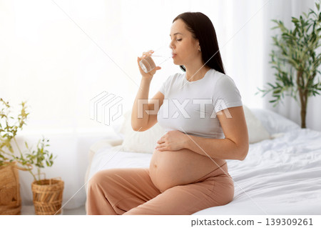 Pregnant woman sitting on bed drinking glass of water while cradling belly Pregnant woman sitting on bed drinking glass of water while cradling belly 139309261