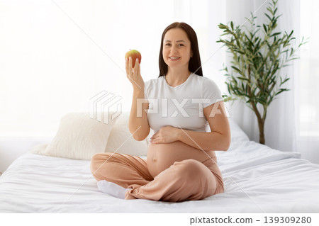 Smiling pregnant woman sitting on bed holding red apple and touching belly Smiling pregnant woman sitting on bed holding red apple and touching belly 139309280