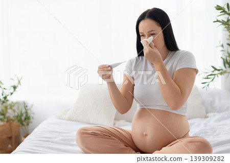 Sick pregnant woman on bed blowing nose into tissue and holding thermometer Sick pregnant woman on bed blowing nose into tissue and holding thermometer 139309282
