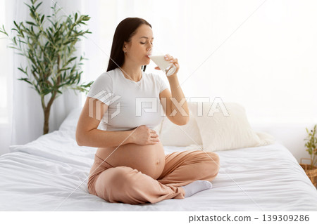 Pregnant woman sitting on bed drinking glass of milk while cradling bare belly Pregnant woman sitting on bed drinking glass of milk while cradling bare belly 139309286