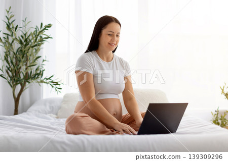 Smiling pregnant woman sitting on bed using laptop, browsing or working online 139309296