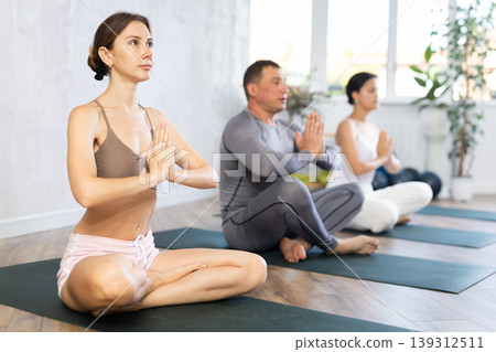 Young beautiful girl with other people relaxing and enjoying yoga in fitness center, meditation in lotus pose Young beautiful girl with other people relaxing and enjoying yoga in fitness center, meditation in lotus pose 139312511