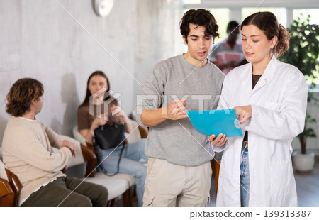 Young female nurse or doctor talking with young guy in reception area Young female nurse or doctor talking with young guy in reception area 139313387