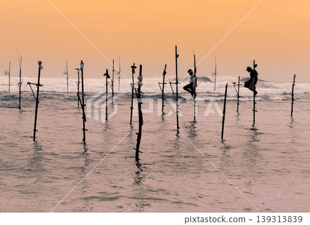 A couple of Stilt fishermen sitting on their poles with wooden fishing rods in their hands in the sunset evening. Ocean waves crash behind them in the background with an orange yellowish sky. A couple of Stilt fishermen sitting on their poles with wooden fishing rods in their hands in the sunset evening. Ocean waves crash behind them in the background with an orange yellowish sky. 139313839