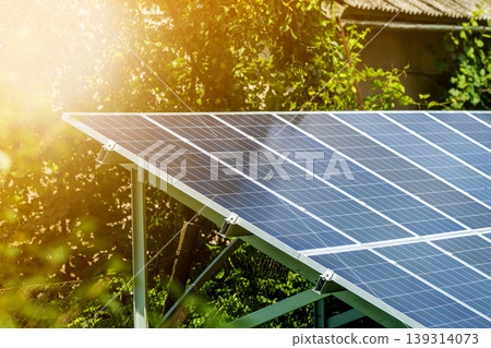Close-up surface of lit by sun modern saving efficient stand -alone blue shiny solar photo voltaic panels system producing renewable clean energy on green grass and trees foliage background. Close-up surface of lit by sun modern saving efficient stand -alone blue shiny solar photo voltaic panels system producing renewable clean energy on green grass and trees foliage background. 139314073
