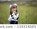 Close-up portrait of cute adorable smiling confident first grader girl in school uniform and white bows in long blond hair on blurred light green sunny outdoors copy space background. 139314085