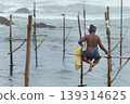 Stilt fisherman with his wooden rod facing back to the camera without a top, fishing in a traditional unique method in Sri Lankan culture, sunny bright evening on the beach. 139314625
