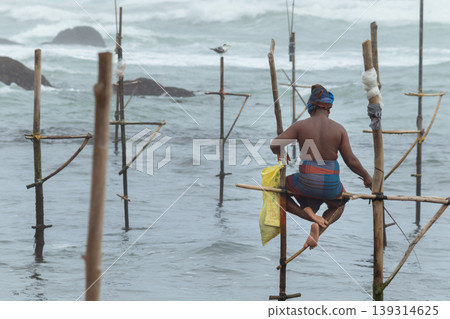 Stilt fisherman with his wooden rod facing back to the camera without a top, fishing in a traditional unique method in Sri Lankan culture, sunny bright evening on the beach. 139314625