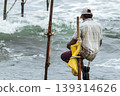 Stilt fisherman with his wooden rod facing back to the camera with his yellow pocket on the pole, fishing in a traditional unique method in Sri Lankan culture, sunny bright evening on the beach. 139314626