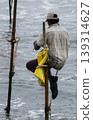 Stilt fisherman with his wooden rod facing back to the camera with his yellow pocket on the pole, fishing in a traditional unique method in Sri Lankan culture, sunny bright evening on the beach. 139314627