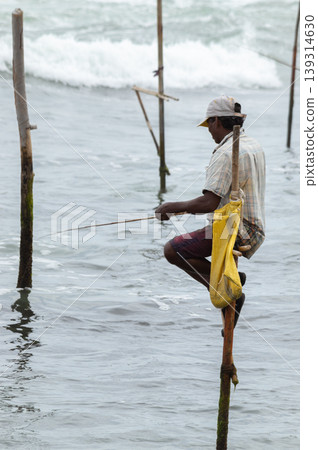 Stilt fisherman with his wooden rod facing side to the camera with his yellow pocket on the pole, fishing in a traditional unique method in Sri Lankan culture, sunny bright evening on the beach. Stilt fisherman with his wooden rod facing side to the camera with his yellow pocket on the pole, fishing in a traditional unique method in Sri Lankan culture, sunny bright evening on the beach. 139314630