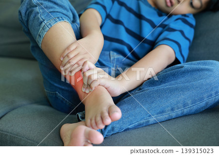 Asian little boy suffering from ankle pain holding injured leg with red glowing spot sitting on grey sofa at home. Sick child having muscle cramp, sprain, or bone injury. Pediatric healthcare. Asian little boy suffering from ankle pain holding injured leg with red glowing spot sitting on grey sofa at home. Sick child having muscle cramp, sprain, or bone injury. Pediatric healthcare. 139315023