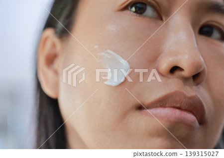 Smiling beautiful Asian woman applying white moisturizing face cream on her cheek with her finger. Daily skincare beauty routine concept for healthy glowing skin, anti-aging and facial hydration. Smiling beautiful Asian woman applying white moisturizing face cream on her cheek with her finger. Daily skincare beauty routine concept for healthy glowing skin, anti-aging and facial hydration. 139315027