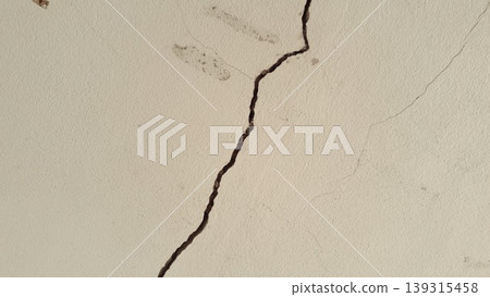 Close-up of a deep, irregular crack running through an aged, textured white concrete wall with minor surface damage. 139315458