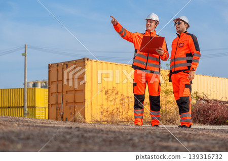 Caucasian Male Engineers in Orange Safety Uniforms Pointing at Shipping Containers in Europe Logistics Yard Caucasian Male Engineers in Orange Safety Uniforms Pointing at Shipping Containers in Europe Logistics Yard 139316732