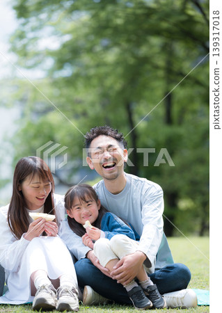 A family enjoying lunch in a park surrounded by fresh greenery, their smiles looking directly at the camera. Perfect for holidays, outings, and leisure. A family enjoying lunch in a park surrounded by fresh greenery, their smiles looking directly at the camera. Perfect for holidays, outings, and leisure. 139317018