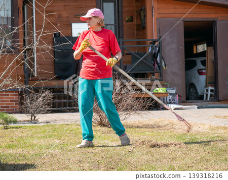 Woman gardener raking dry grass in backyard. Mature lady cleaning lawn with rake during spring season. Seasonal outdoor property maintenance, house chore and landscaping lifestyle activity. 139318216