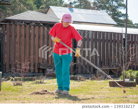 Woman gardener using rake to clean lawn in spring. Backyard housekeeping with outdoor tool. Senior female working in garden during sunny day. Horticulture and seasonal yard maintenance. Woman gardener using rake to clean lawn in spring. Backyard housekeeping with outdoor tool. Senior female working in garden during sunny day. Horticulture and seasonal yard maintenance. 139318217