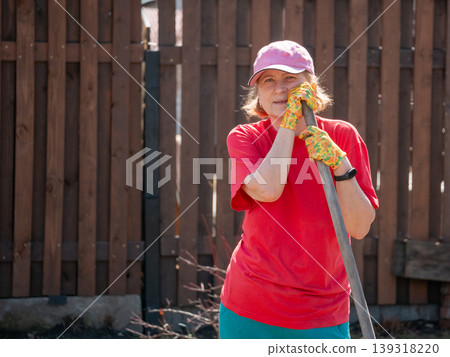 Senior woman gardener leaning on spade in backyard. Mature lady wearing cap and work glove doing yard work. Hobby of horticulture and vegetable growing in spring season near house fence. Senior woman gardener leaning on spade in backyard. Mature lady wearing cap and work glove doing yard work. Hobby of horticulture and vegetable growing in spring season near house fence. 139318220