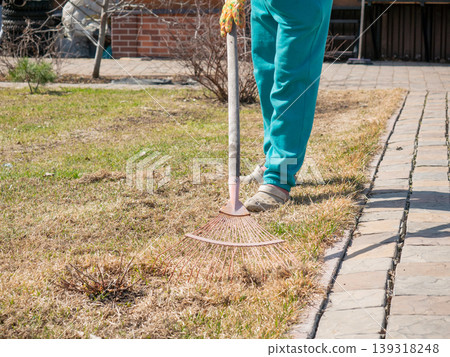 Woman raking dry grass in garden during spring cleaning. Gardener clearing lawn with metal rake. Seasonal yard maintenance work. Agriculture hobby and house land improvement concept. Woman raking dry grass in garden during spring cleaning. Gardener clearing lawn with metal rake. Seasonal yard maintenance work. Agriculture hobby and house land improvement concept. 139318248