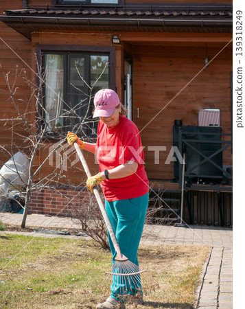 Woman gardener using rake to clean lawn in backyard. Senior woman doing yard maintenance at home. Spring garden work, landscaping, hobby, lifestyle activity, seasonal outdoor chore. 139318249