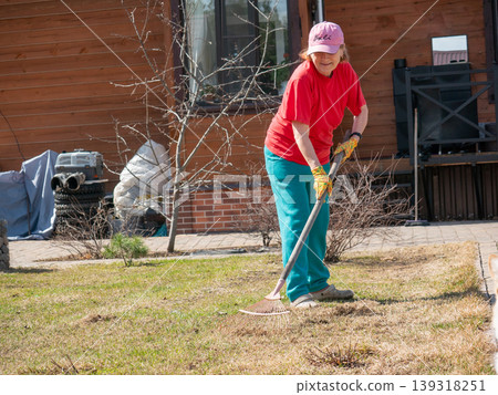 Senior woman gardener raking dry grass in backyard. Elderly lady cleaning lawn with rake in spring. Maintenance of home garden, seasonal outdoor work, healthy active lifestyle for senior. 139318251
