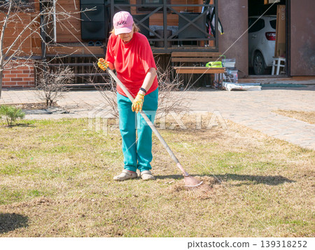 Senior woman raking dry grass in a home lawn. Gardener working in the backyard during spring season. Seasonal yard maintenance, cleaning garden soil, and landscape care activity concept. Senior woman raking dry grass in a home lawn. Gardener working in the backyard during spring season. Seasonal yard maintenance, cleaning garden soil, and landscape care activity concept. 139318252
