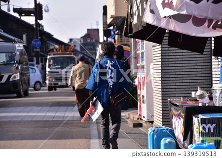 Inuyama City, Aichi Prefecture: A man participating in the Inuyama Festival, walking through the castle town wearing a happi coat with "Inuyama Festival" written on it, and stalls being prepared to open. Inuyama City, Aichi Prefecture: A man participating in the Inuyama Festival, walking through the castle town wearing a happi coat with "Inuyama Festival" written on it, and stalls being prepared to open. 139319513