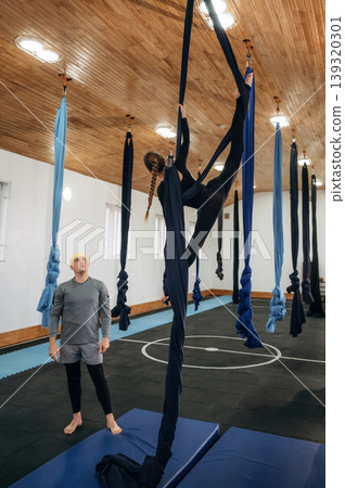 In the air, high up. Male instructor assisting a child in aerial yoga practice on hammock straps in a fitness studio In the air, high up. Male instructor assisting a child in aerial yoga practice on hammock straps in a fitness studio 139320301