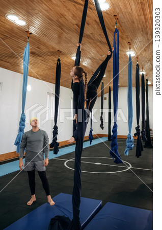 Looking and controlling. Male instructor assisting a child in aerial yoga practice on hammock straps in a fitness studio Looking and controlling. Male instructor assisting a child in aerial yoga practice on hammock straps in a fitness studio 139320303