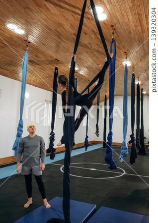 Looking and controlling. Male instructor assisting a child in aerial yoga practice on hammock straps in a fitness studio Looking and controlling. Male instructor assisting a child in aerial yoga practice on hammock straps in a fitness studio 139320304
