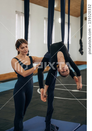 Private session, hanging. Female instructor helping a child practice aerial yoga on hammock straps in a fitness studio Private session, hanging. Female instructor helping a child practice aerial yoga on hammock straps in a fitness studio 139320332