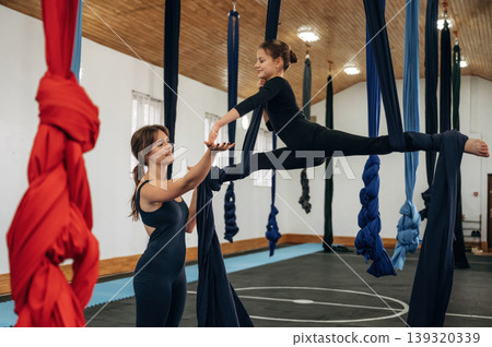 Two people, sport. Female instructor helping a child practice aerial yoga on hammock straps in a fitness studio Two people, sport. Female instructor helping a child practice aerial yoga on hammock straps in a fitness studio 139320339