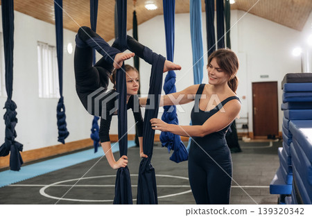 With legs high up. Female instructor helping a child practice aerial yoga on hammock straps in a fitness studio 139320342