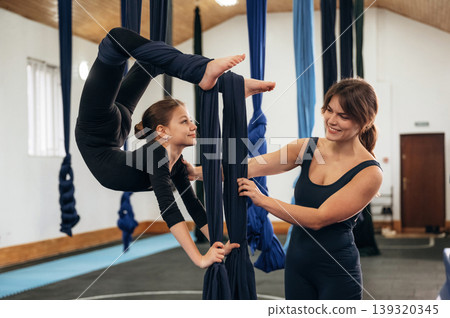 Cooperating with the girl. Female instructor helping a child practice aerial yoga on hammock straps in a fitness studio 139320345