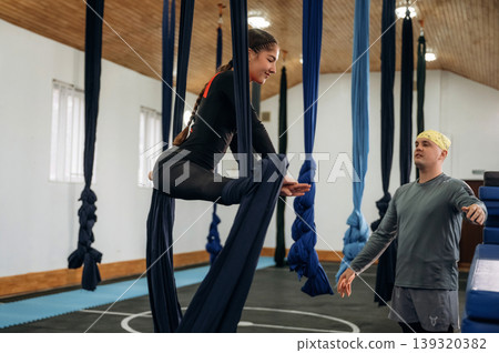 Girl is on hammock straps. Male instructor assisting a child in aerial yoga practice in a fitness studio Girl is on hammock straps. Male instructor assisting a child in aerial yoga practice in a fitness studio 139320382