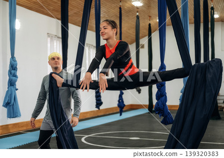 Girl is on hammock straps. Male instructor assisting a child in aerial yoga practice in a fitness studio Girl is on hammock straps. Male instructor assisting a child in aerial yoga practice in a fitness studio 139320383