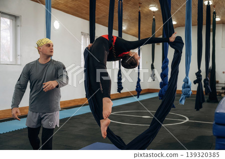 Girl is on hammock straps. Male instructor assisting a child in aerial yoga practice in a fitness studio Girl is on hammock straps. Male instructor assisting a child in aerial yoga practice in a fitness studio 139320385