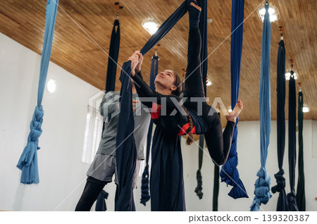 Male instructor assisting a child in aerial yoga practice on hammock straps in a fitness studio Male instructor assisting a child in aerial yoga practice on hammock straps in a fitness studio 139320387