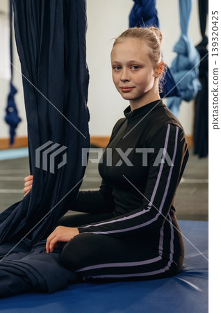 Looking at the camera, sitting on the mat. Child practicing aerial yoga on hammock straps in a fitness studio Looking at the camera, sitting on the mat. Child practicing aerial yoga on hammock straps in a fitness studio 139320425