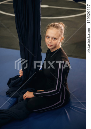 Looking at the camera, sitting on the mat. Child practicing aerial yoga on hammock straps in a fitness studio 139320427
